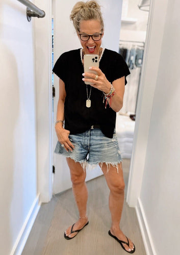 A woman stands in front of a mirror, taking a selfie with her phone. She is smiling and wearing glasses, a black shirt, Moussy Packard Shorts with frayed edges and button fly, flip-flops, and several bracelets. Her hair is tied up, and she is in a light-colored room.