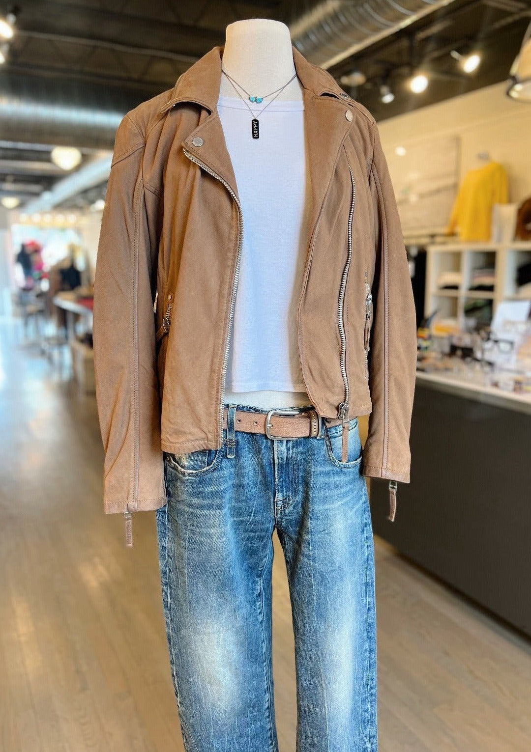 A mannequin dressed in a Nubuck Jacket by Mauritius over a Perfect White Tee, paired with R13 jeans and a beige belt. The outfit is displayed in a well-lit, modern clothing store with racks of clothes and shelves in the background.