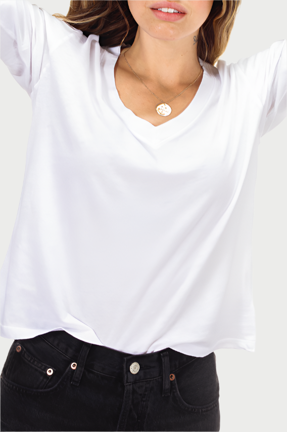 A woman with wavy brown hair is standing against a plain background. She is wearing a perfectwhitetee Jimi Long Sleeve V Neck Tee partially tucked into dark pants. With her arms raised and hands behind her head, she showcases a necklace and bracelets while smiling slightly.