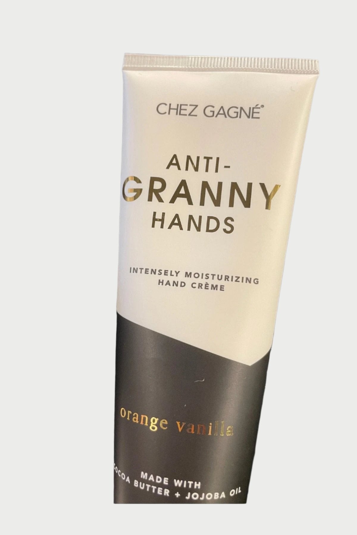 A tube of Chez Gagné Hand Creme with orange and vanilla scent lies on a dark, textured fabric. The packaging highlights its rich blend of cocoa butter and jojoba oil.