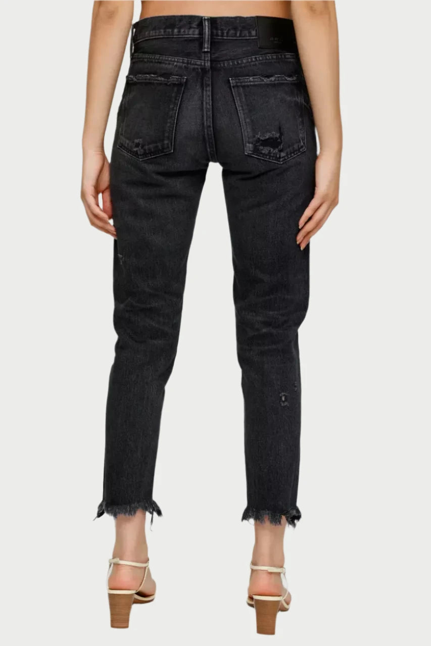 A person wearing the Merry Tapered Mid Rise Jeans by Moussy, crafted from Japanese denim with a distressed black finish, button fly, and fitted waistband, stands with their back to the camera. The jeans have frayed hems and are paired elegantly with beige heeled sandals.