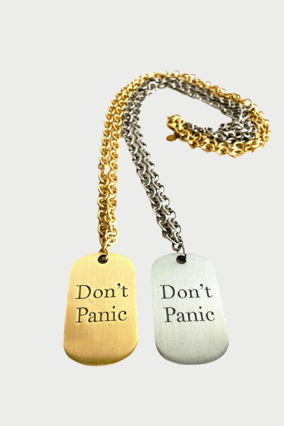 Two hand-crafted Don't Panic Dog Tag Necklace by Simply Eartha with the inscription "Don't Panic" lie on a white surface. One tag features an antique finish and a matching gold chain, and the other tag is silver with a heavy gauge chain. The chains are partially intertwined.