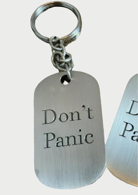 Close-up of a vintage silver brass keychain with an engraved rectangular tag. The tag has the text "Don't Panic" etched onto it. The background appears textured with a rough, metallic surface, reminiscent of a Simply Eartha Don’t Panic key chain. Another similar keychain is partially visible on the right.