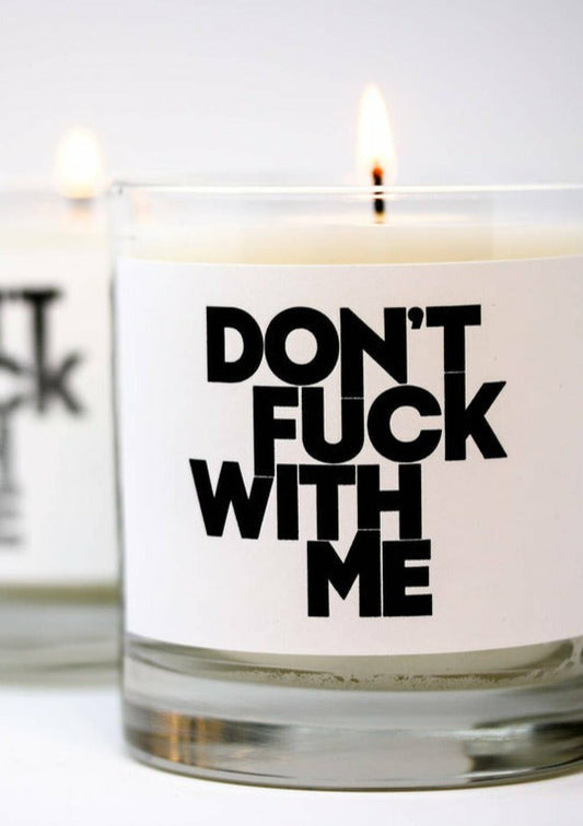 A clear glass Thompson Ferrier Candle with a white label containing the bold, black text "DON'T FUCK WITH ME." The candle by Thompson Ferrier, made from a soy wax blend, is lit, and another similar candle is blurred in the background.