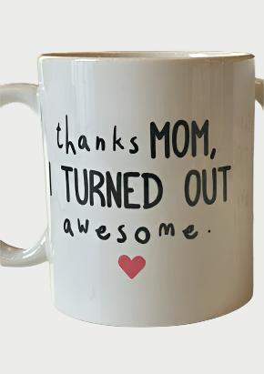 A white ceramic Thanks Mom Mug from 365 Printing Inc with the text "thanks MOM, I TURNED OUT awesome." printed on it in a playful font. There is a small red heart underneath the text, making it the perfect Mother's gift. This 11oz mug adds a touch of love to your morning coffee.