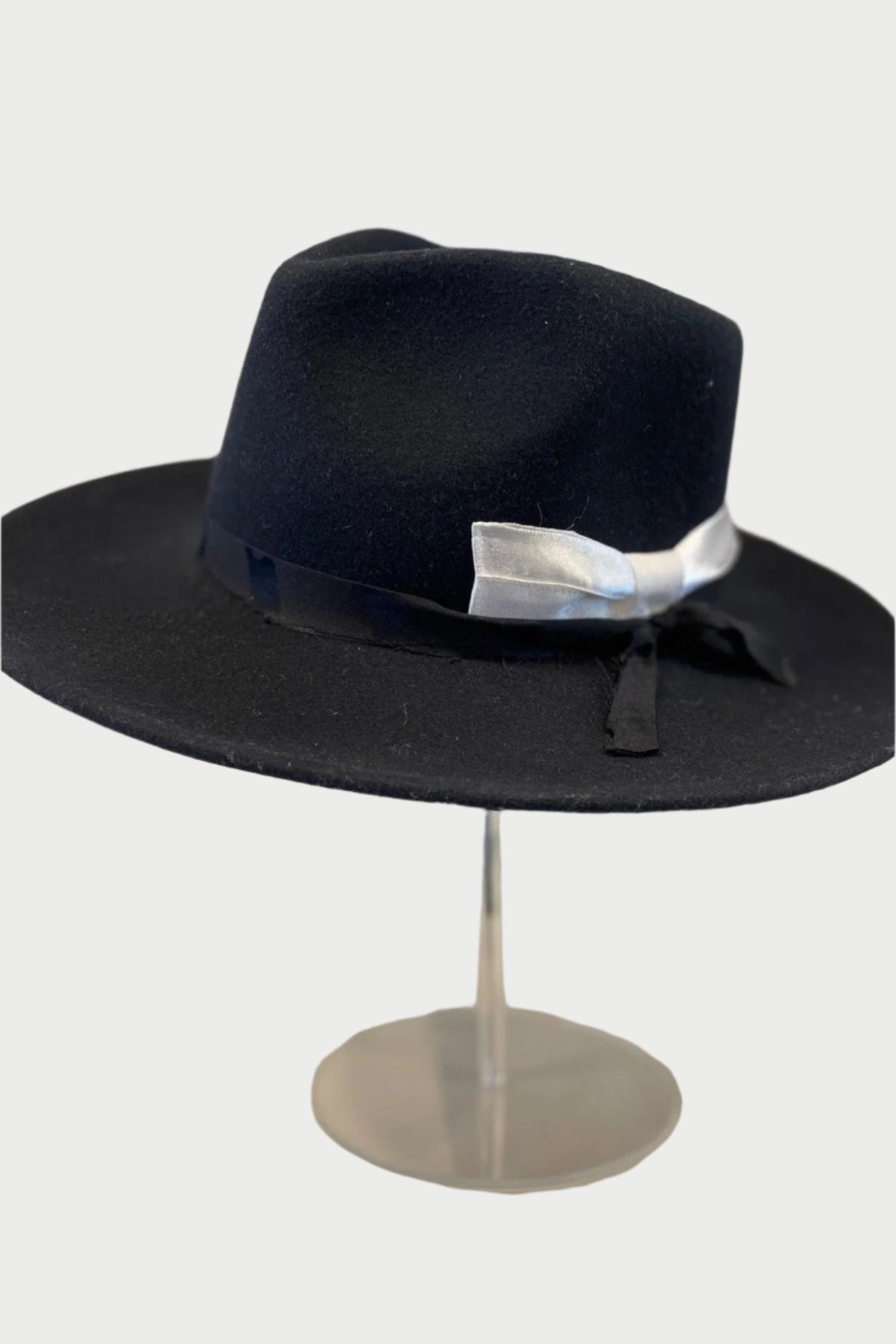 A dark blue wide-brimmed hat, made from up-cycled material, is displayed on a clear stand. The Upcycled Hats by Label Jae feature a black ribbon adorned with a small white knot on one side. The background is neutral and slightly blurred, emphasizing the hat as the main focus.