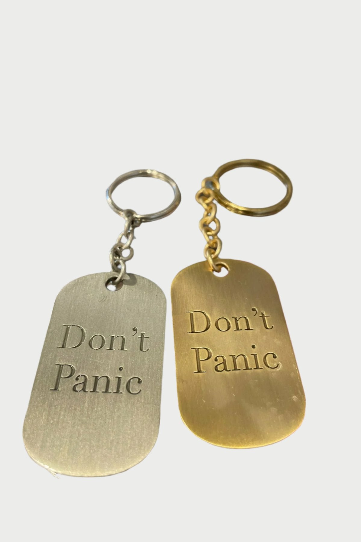 Two keychain dog tags are displayed on a glass surface. Both are rectangular with rounded edges, resembling vintage designs. One is silver brass with the inscription "Don't Panic" in black, and the other is gold with the same inscription in green. These are part of the Don’t Panic key chain collection by Simply Eartha.